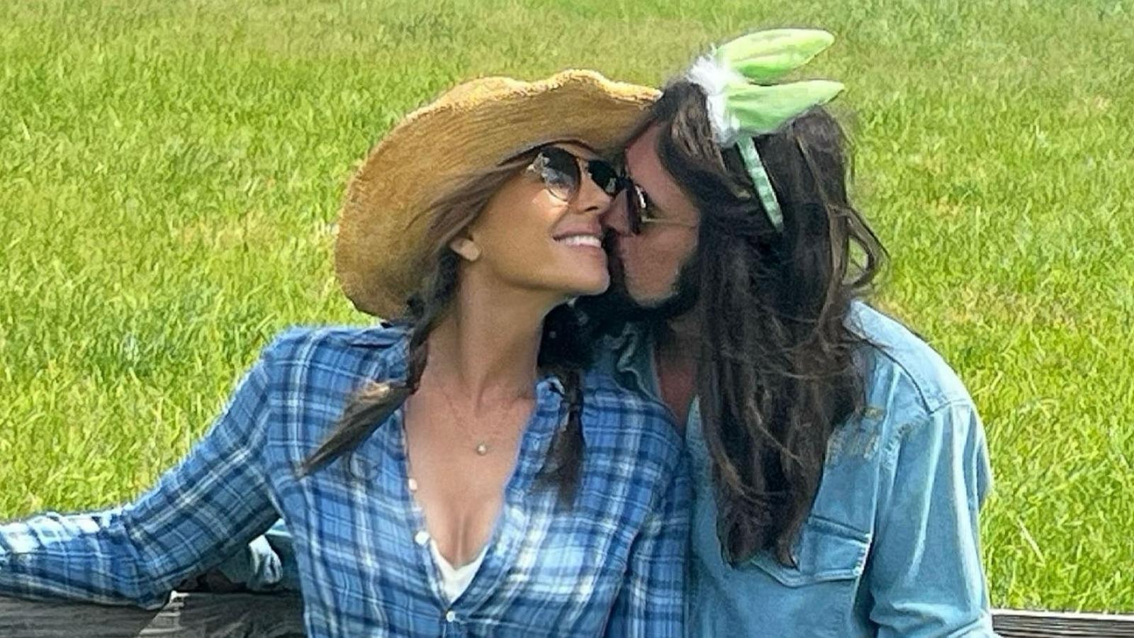 Elizabeth Hurley in plaid shirt and cowboy hat as Billy Ray Cyrus, wearing green bunny ears, kisses her cheek beside a wooden fence